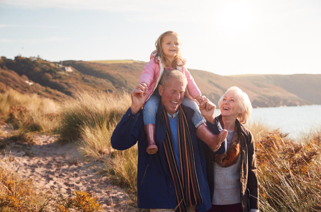 Grandfather,Giving,Granddaughter,Ride,On,Shoulders,As,They,Walk,Through - Gardner Physical Therapy Grandparents walking on the beach with their granddaughter, representing improved stability and independence through balance training at Gardner Physical Therapy.”
