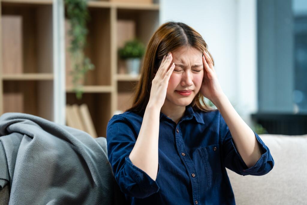 A,Young,Asian,Woman,Sits,On,A,Sofa,In,Her - Gardner Physical Therapy Young woman holding her head in pain, showing common concussion or post-concussion symptoms.