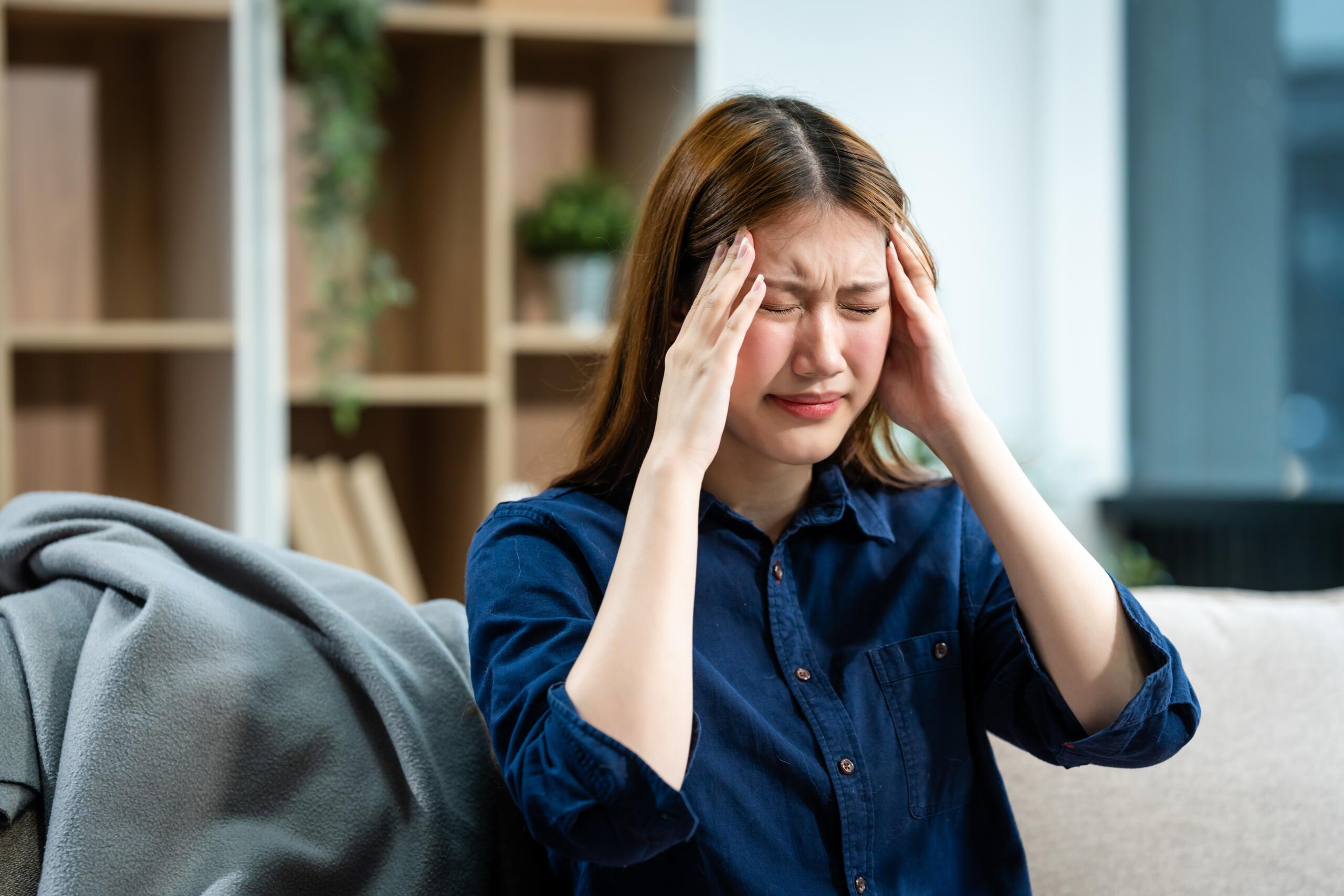 A,Young,Asian,Woman,Sits,On,A,Sofa,In,Her - Gardner Physical Therapy Young woman holding her head in pain, showing common concussion or post-concussion symptoms.