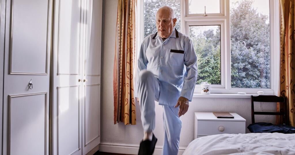 Fitness,,Bedroom,And,Senior,Man,Lifting,Legs,In,A,Nursing - Gardner Physical Therapy Senior man practicing balance exercises at home to improve stability and reduce fall risk with Gardner Physical Therapy’s fall prevention program.