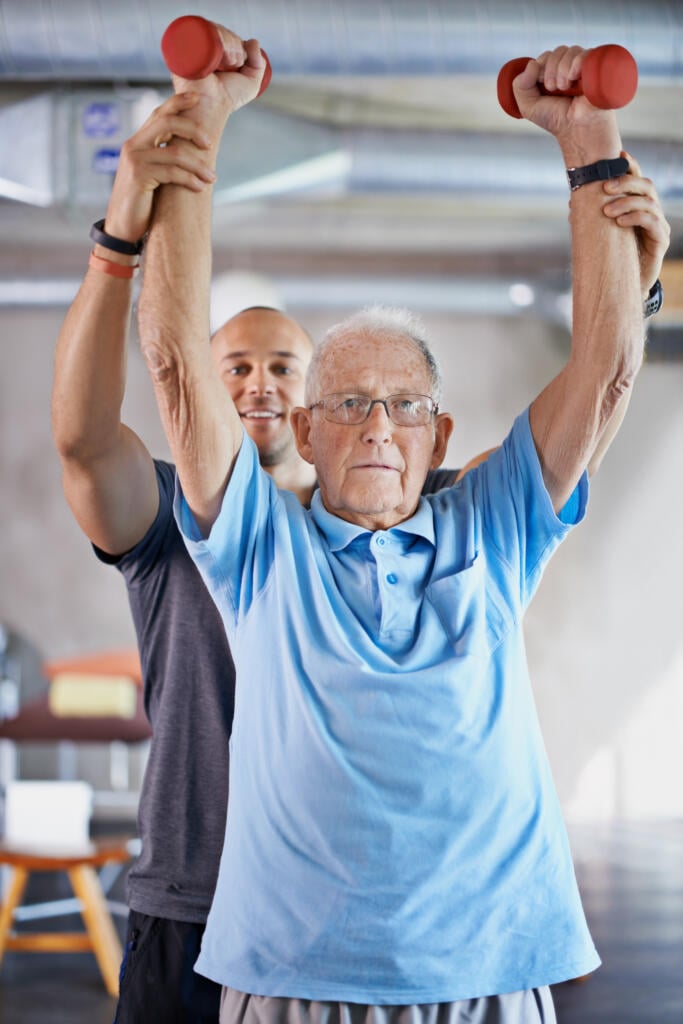Senior man lifting light dumbbells overhead with assistance from a physical therapist during a rehabilitation session.