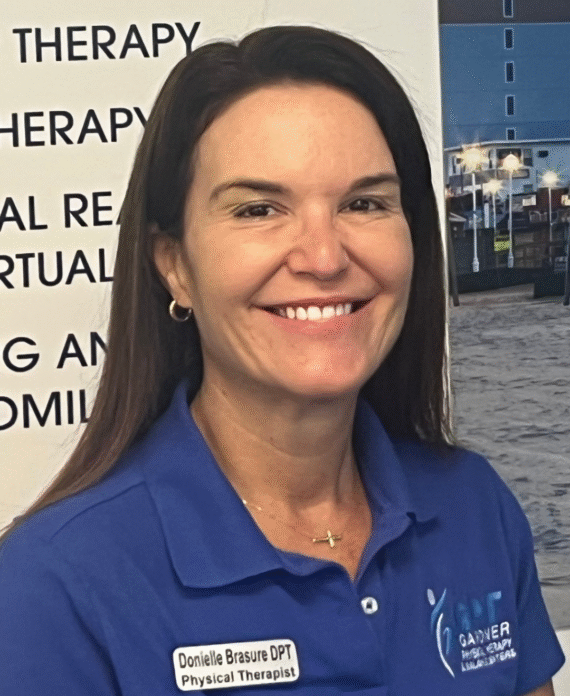 Dr. Donielle Brasure, DPT, NCS, smiling at Gardner Physical Therapy’s Ocean Pines location, standing in front of the clinic’s vestibular and neurologic services display.