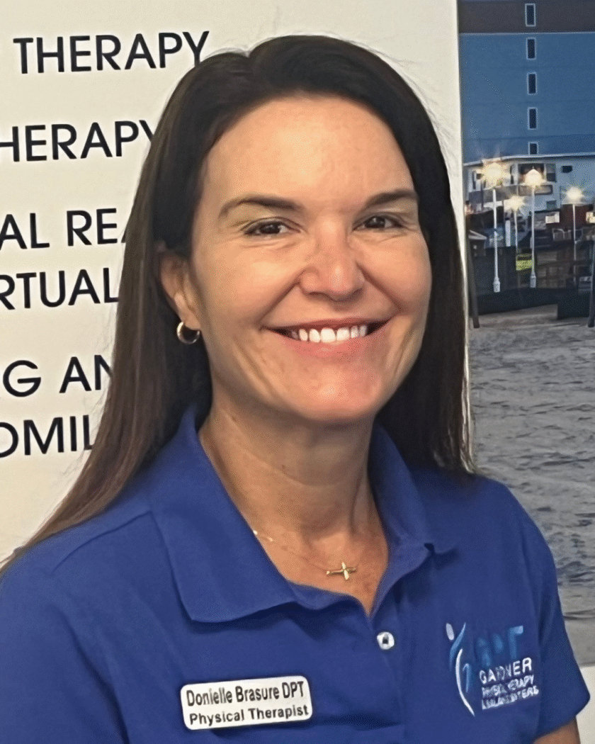 Dr. Donielle Brasure, DPT, NCS, smiling at Gardner Physical Therapy’s Ocean Pines location, standing in front of the clinic’s vestibular and neurologic services display.