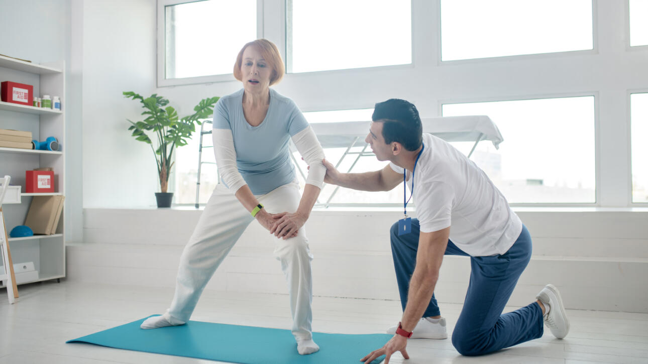Senior woman stretching on a yoga mat with therapist’s support during Gardner Physical Therapy session.