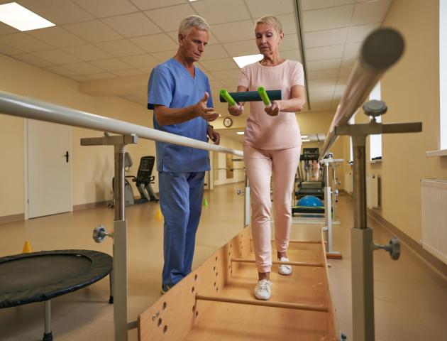 Physiotherapist,Helping,Mature,Patient,Walk,Between,Parallel,Bars,In,Hospital